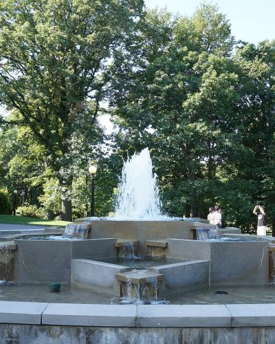 A multi-tiered stone fountain with water jets, situated on a paved terrace with a large stone building and a flagpole visible to the left and dense trees in the background.