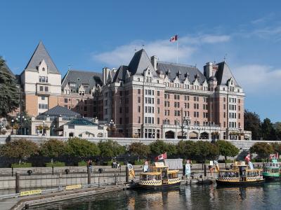 A large, historic Châteauesque-style hotel with pinkish-red brick and grey slate roofs, situated on a waterfront with tour boats docked in the foreground and a Canadian flag flying from the roof.
