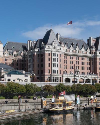 A large, historic Châteauesque-style hotel with pinkish-red brick and grey slate roofs, situated on a waterfront with tour boats docked in the foreground and a Canadian flag flying from the roof.