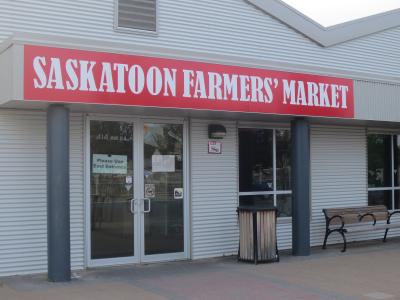 The image shows the exterior entrance of a building with a large red sign reading "SASKATOON FARMERS' MARKET" above glass doors, with the address "414 AVE B S" visible on the window.