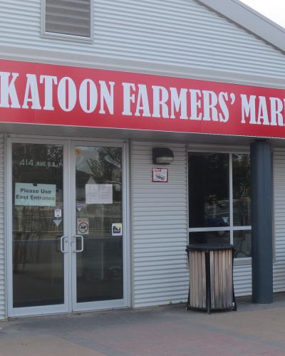 The image shows the exterior entrance of a building with a large red sign reading "SASKATOON FARMERS' MARKET" above glass doors, with the address "414 AVE B S" visible on the window.