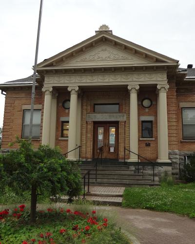 A red brick building with a classical portico featuring four columns and a pediment, with the text "PUBLIC LIBRARY" clearly visible above the entrance doors.