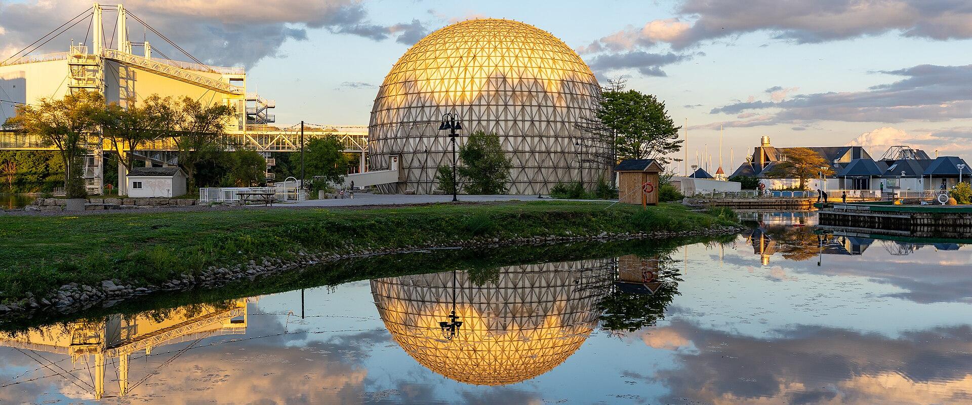 A large geodesic dome structure (the Cinesphere) and a white exhibition hall (the Ontario Place Exhibition Hall) situated on a waterfront with their reflections visible in the water.