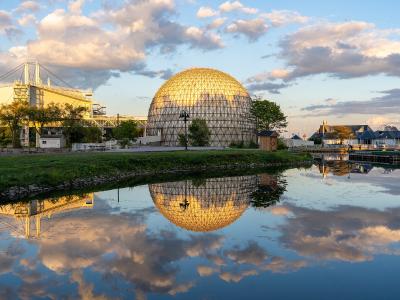 A large geodesic dome structure (the Cinesphere) and a white exhibition hall (the Ontario Place Exhibition Hall) situated on a waterfront with their reflections visible in the water.
