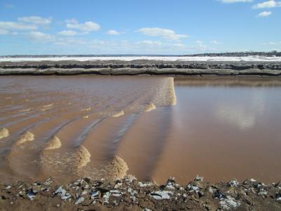 A muddy tidal bore wave moving upstream in a wide river channel with snow-covered banks and a distant shoreline under a blue sky.