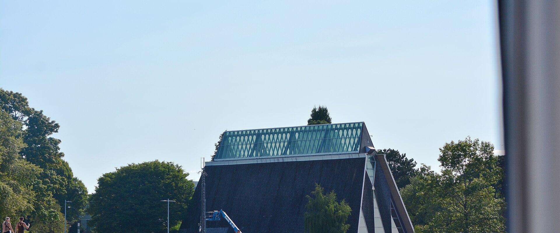 A distinctive A-frame building with a dark shingled facade and a large glass skylight roof, situated on a rocky shoreline with trees and people nearby.