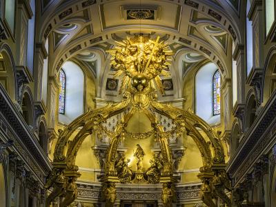 The image shows the ornate interior of a cathedral featuring a massive golden baldachin (canopy) over the main altar, with Baroque architectural details, statues, and a painting of the Virgin Mary.