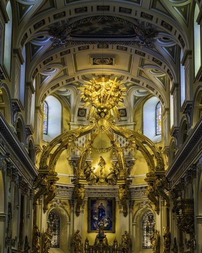 The image shows the ornate interior of a cathedral featuring a massive golden baldachin (canopy) over the main altar, with Baroque architectural details, statues, and a painting of the Virgin Mary.