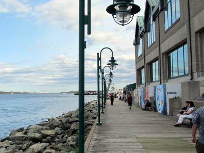 A wooden boardwalk path lined with distinctive green curved lampposts runs alongside a rocky harbor edge, with a large multi-story building on the right and people walking or sitting along the path.