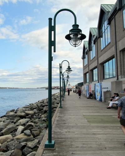 A wooden boardwalk path lined with distinctive green curved lampposts runs alongside a rocky harbor edge, with a large multi-story building on the right and people walking or sitting along the path.