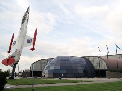A large white and red jet aircraft mounted vertically on a pedestal stands in front of a modern building featuring a distinctive dark glass geodesic dome and arched hangar wings.