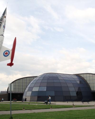 A large white and red jet aircraft mounted vertically on a pedestal stands in front of a modern building featuring a distinctive dark glass geodesic dome and arched hangar wings.