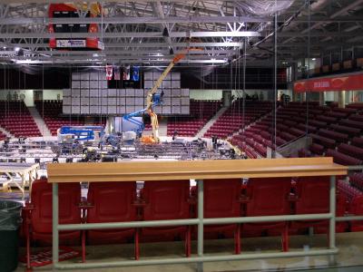 Interior view of a large indoor sports arena with red stadium seating, a suspended scoreboard, and an ice rink surface under construction or maintenance using lifts and scaffolding.