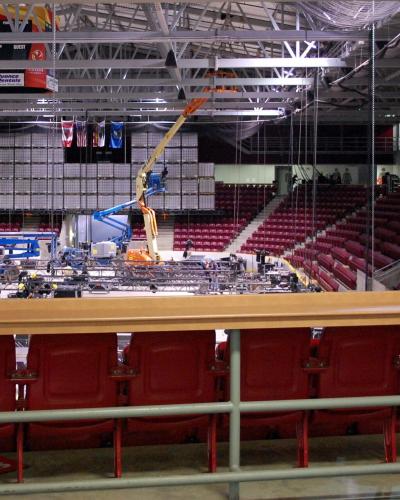 Interior view of a large indoor sports arena with red stadium seating, a suspended scoreboard, and an ice rink surface under construction or maintenance using lifts and scaffolding.