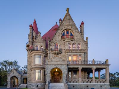 A large, ornate stone castle with a red roof, multiple turrets, and balconies, photographed at twilight with lights on inside.