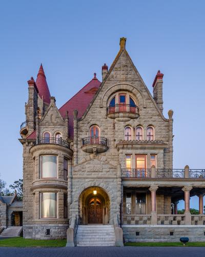 A large, ornate stone castle with a red roof, multiple turrets, and balconies, photographed at twilight with lights on inside.