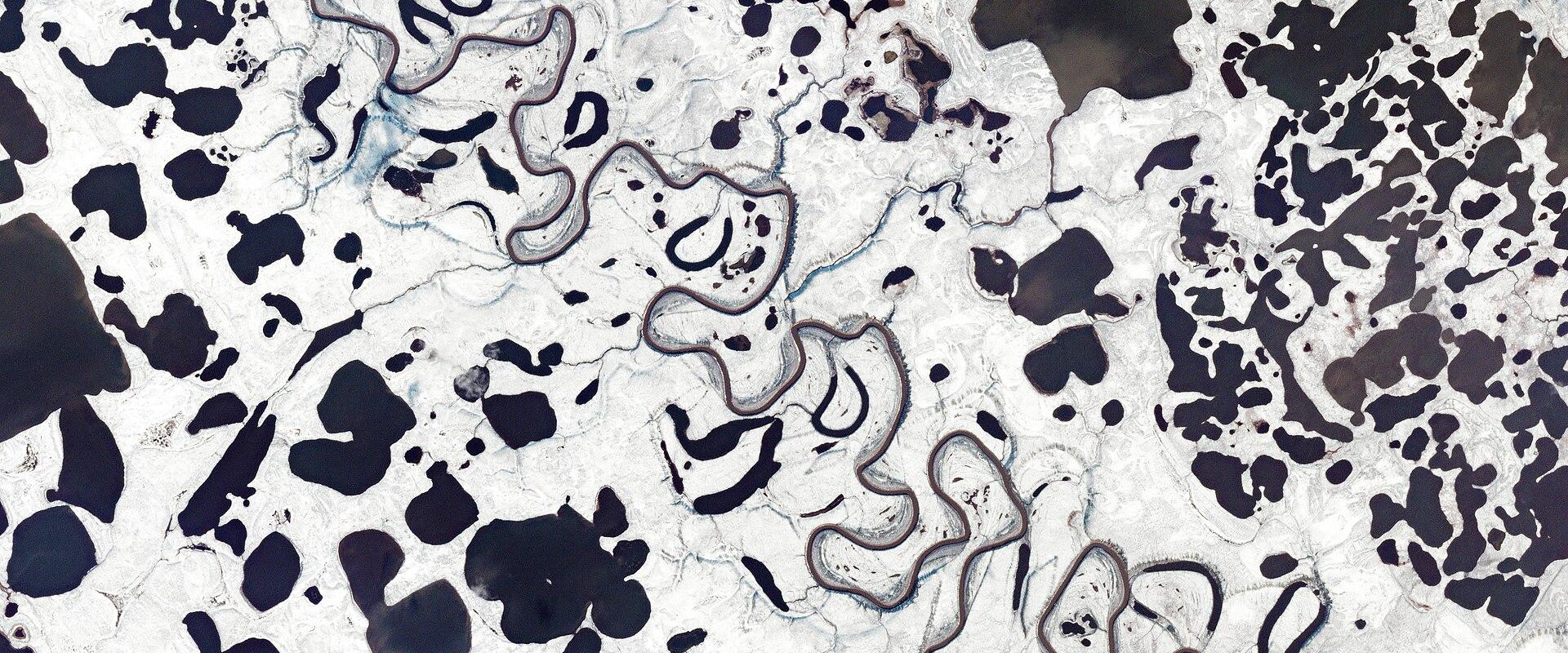 Aerial satellite view of a frozen, snow-covered landscape featuring a meandering river and numerous dark, irregular lakes or ponds.
