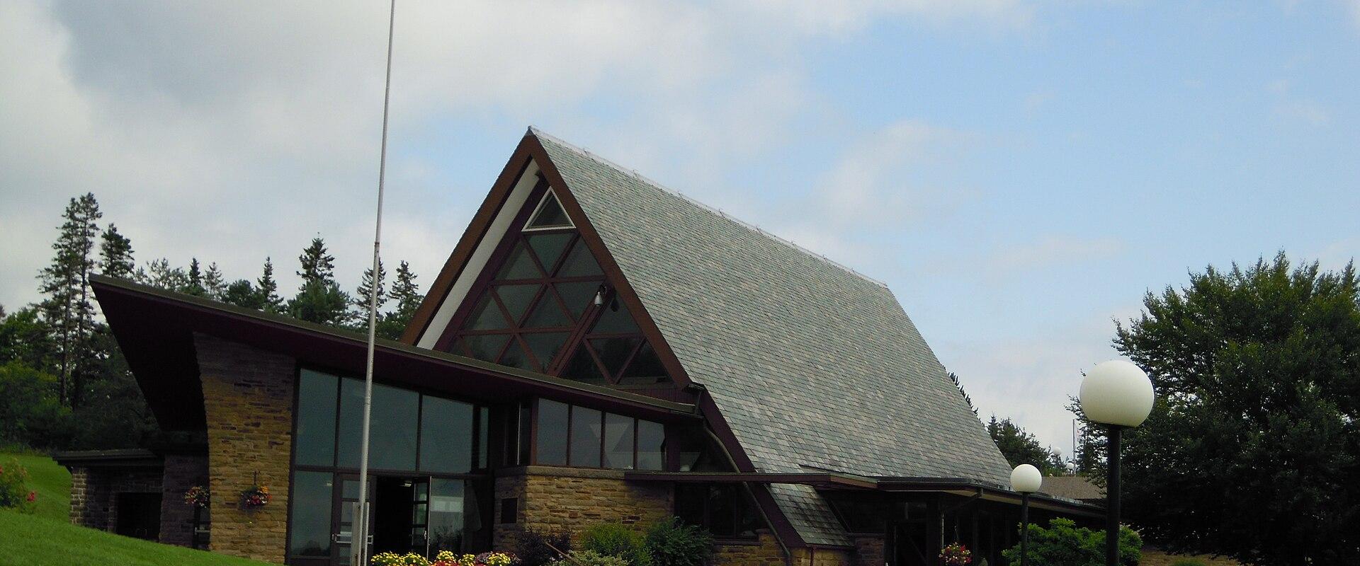 A modern building with a distinctive A-frame roof, stone facade, and large glass windows, situated on a grassy slope with a Canadian flag flying on a tall pole to the left.