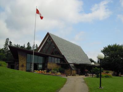 A modern building with a distinctive A-frame roof, stone facade, and large glass windows, situated on a grassy slope with a Canadian flag flying on a tall pole to the left.
