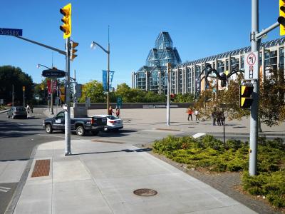 A street-level view of an intersection with a blue street sign reading "Sussex" and a distinctive modern glass building with a pyramid-like roof structure in the background.