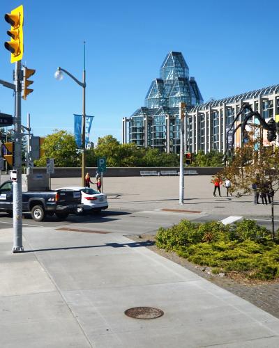A street-level view of an intersection with a blue street sign reading "Sussex" and a distinctive modern glass building with a pyramid-like roof structure in the background.