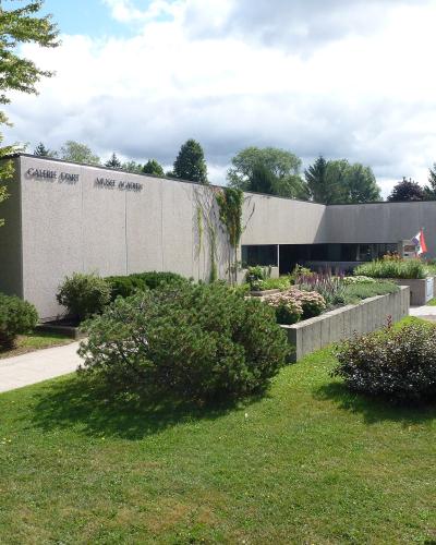 A low-rise concrete building with "GALERIE D'ART MUSÉE ACADIEN" lettering on the facade, surrounded by green lawns and trees.
