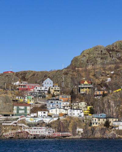 A view of colorful houses built on a steep, rocky hillside overlooking a body of water, with a large rocky peak rising in the background.