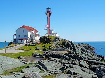 A tall, slender lighthouse tower painted with red and white vertical stripes stands on a rocky headland next to a white keeper's house with a red roof, overlooking the ocean under a clear blue sky.