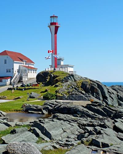 A tall, slender lighthouse tower painted with red and white vertical stripes stands on a rocky headland next to a white keeper's house with a red roof, overlooking the ocean under a clear blue sky.