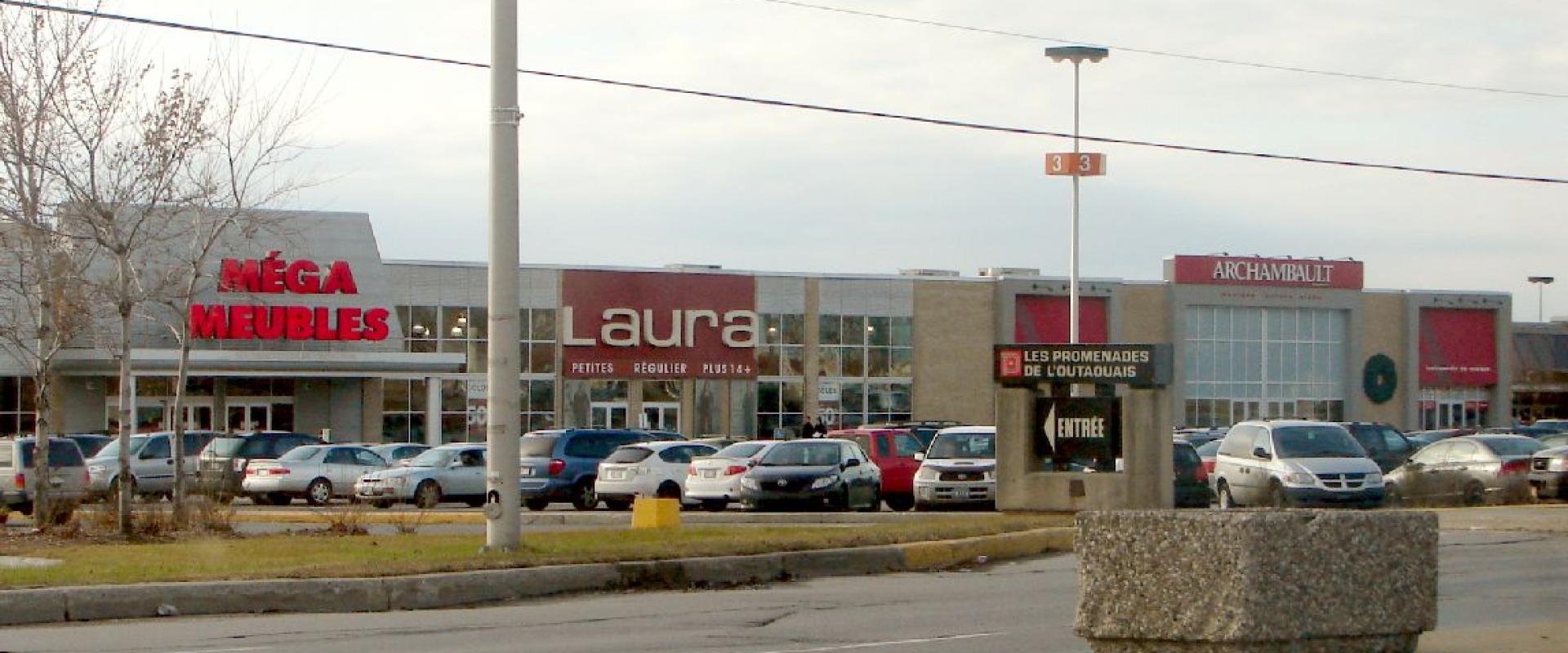 A strip mall shopping center featuring stores like Méga Meubles, Laura, and Archambault, with a sign reading "LES PROMENADES DE L'OUTAOUAIS" and "ENTRÉE".