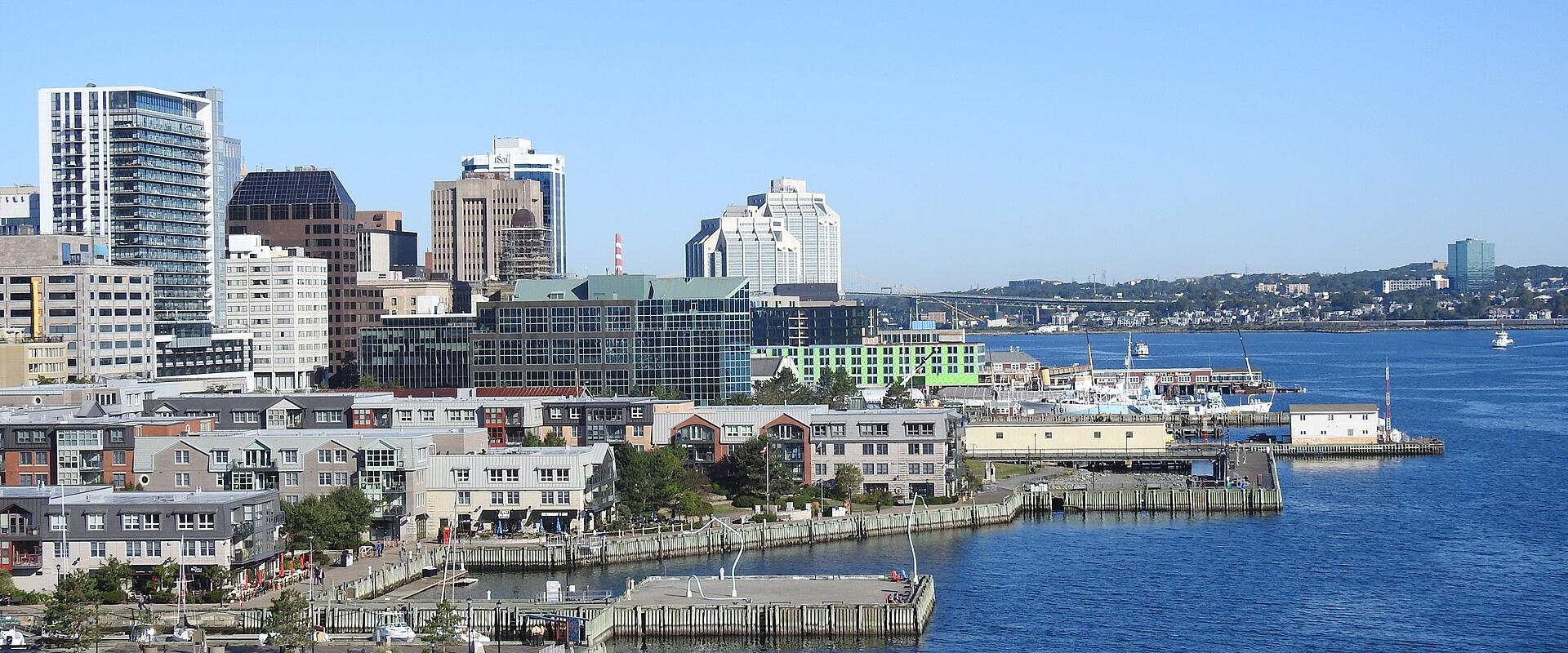 A waterfront view of a city skyline featuring a large construction crane on the left, modern glass buildings, and a row of red-roofed buildings on a pier in the foreground, with the word "MURPHY'S" visible on one of the pier buildings.