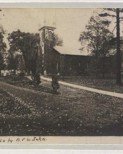 A vintage black and white photograph showing a stone church with a bell tower situated among trees, with a handwritten caption at the bottom reading "St. Mark's Church Niagara-on-the-lake Photo by A.P. St. John."