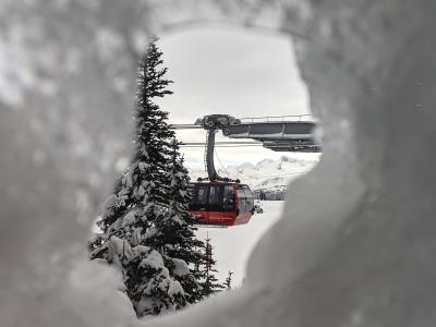 A red gondola cabin is viewed through a snowy foreground, with a support tower and snowy mountain peaks in the background.