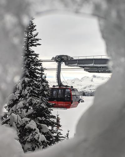A red gondola cabin is viewed through a snowy foreground, with a support tower and snowy mountain peaks in the background.