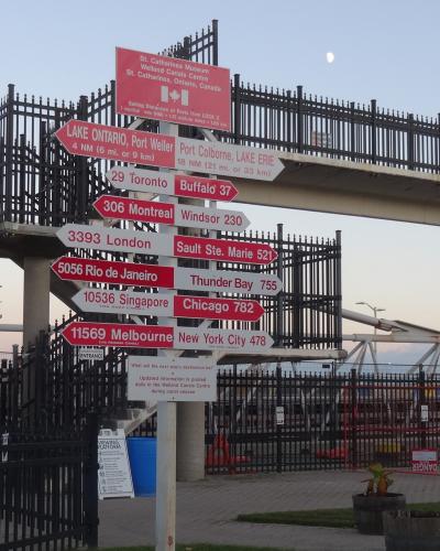 A directional signpost featuring red and white arrows pointing to various global ports and cities, topped with a large red sign reading "St. Catharines Museum Welland Canals Centre St. Catharines, Ontario, Canada".