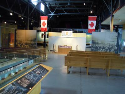The image shows the interior of a museum exhibit featuring rows of wooden benches, a large architectural model of a building in the foreground, and a central display area with a counter labeled "IMMIGRATION" and a sign above it reading "WELCOME TO CA