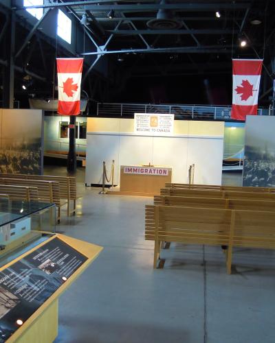 The image shows the interior of a museum exhibit featuring rows of wooden benches, a large architectural model of a building in the foreground, and a central display area with a counter labeled "IMMIGRATION" and a sign above it reading "WELCOME TO CA