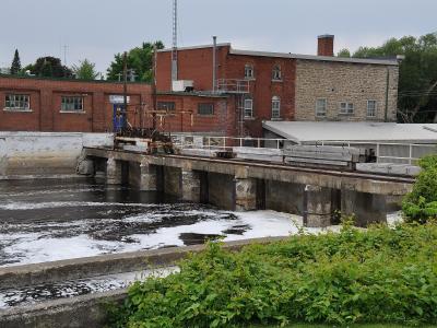 A concrete lock gate structure with a wooden trolley mechanism on rails, situated in a waterway with industrial brick buildings in the background.