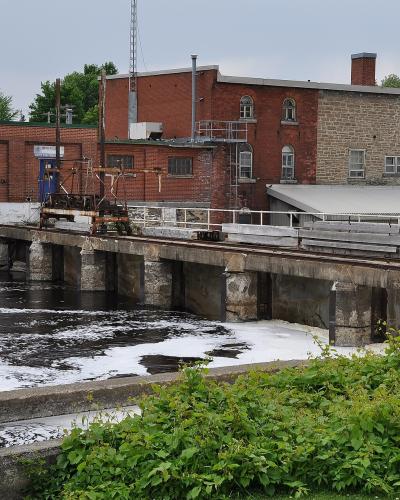 A concrete lock gate structure with a wooden trolley mechanism on rails, situated in a waterway with industrial brick buildings in the background.