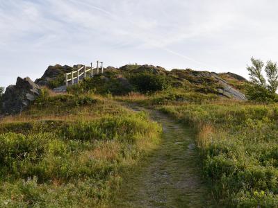 A dirt trail winds up a grassy, rocky hill towards a white wooden fence and viewing platform at the summit.