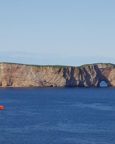 A large, flat-topped limestone sea stack with a prominent natural arch at its base, situated in the ocean with a small red and white boat in the foreground.