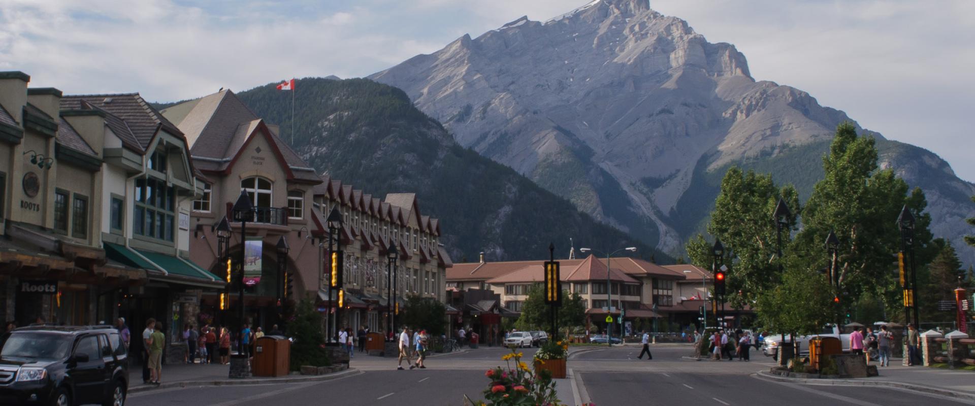 A wooden sign reading "Banff Avenue Square / Place de l'avenue Banff" mounted on a post behind a historical plaque set in rocks within a paved plaza.