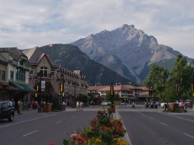 A wooden sign reading "Banff Avenue Square / Place de l'avenue Banff" mounted on a post behind a historical plaque set in rocks within a paved plaza.