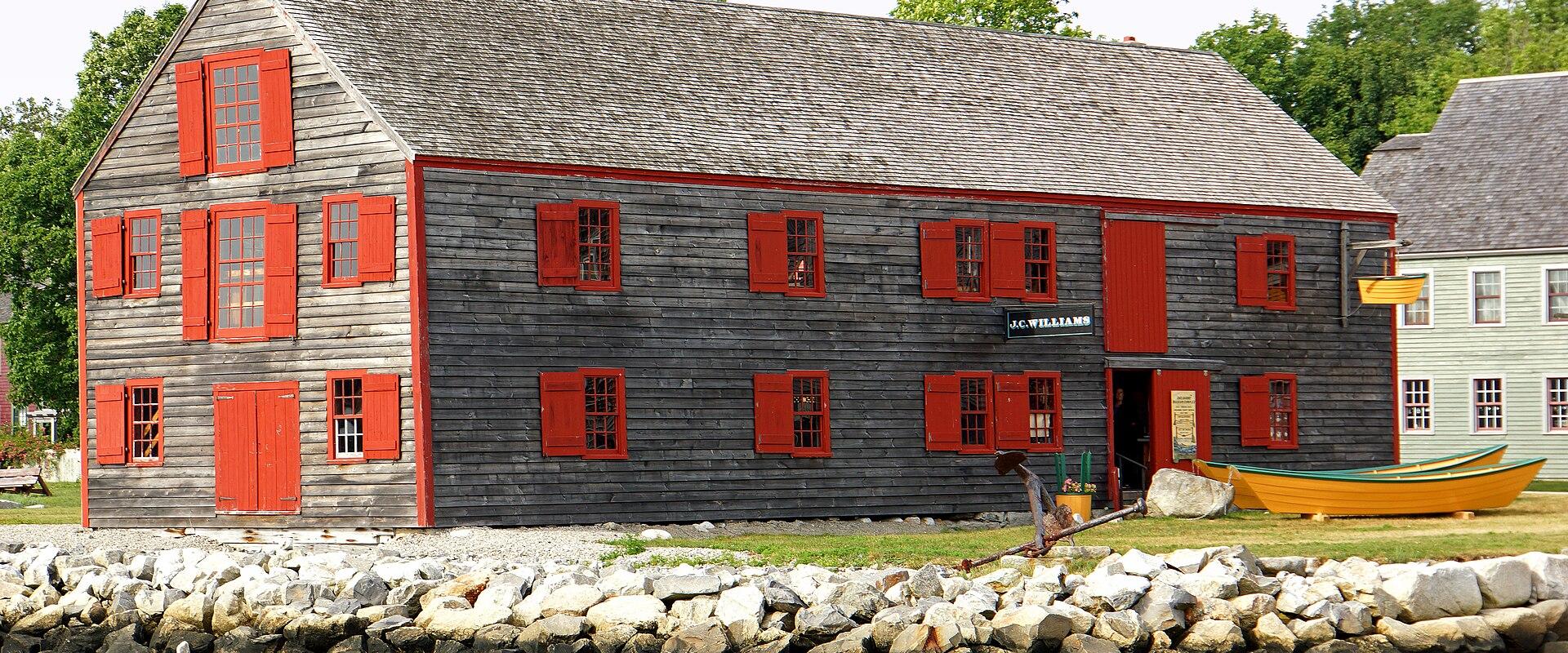 A large, weathered grey wooden building with red trim and shutters, situated on a rocky shore, with a yellow dory boat visible on the grass nearby.