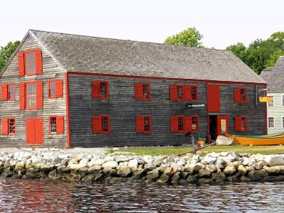 A large, weathered grey wooden building with red trim and shutters, situated on a rocky shore, with a yellow dory boat visible on the grass nearby.