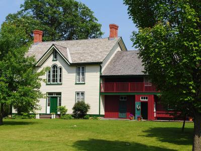 A two-part heritage building complex featuring a white clapboard house with green trim and a red wooden annex with a balcony, set on a grassy lawn with trees.