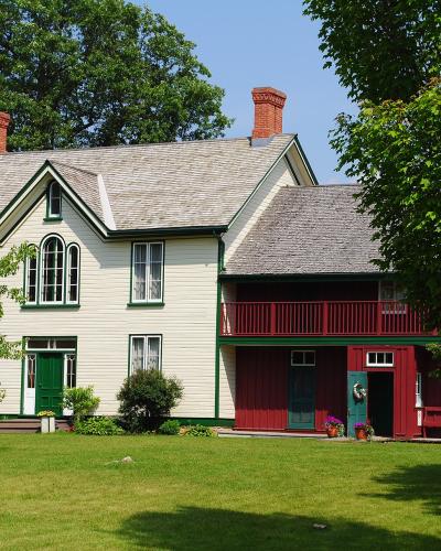 A two-part heritage building complex featuring a white clapboard house with green trim and a red wooden annex with a balcony, set on a grassy lawn with trees.