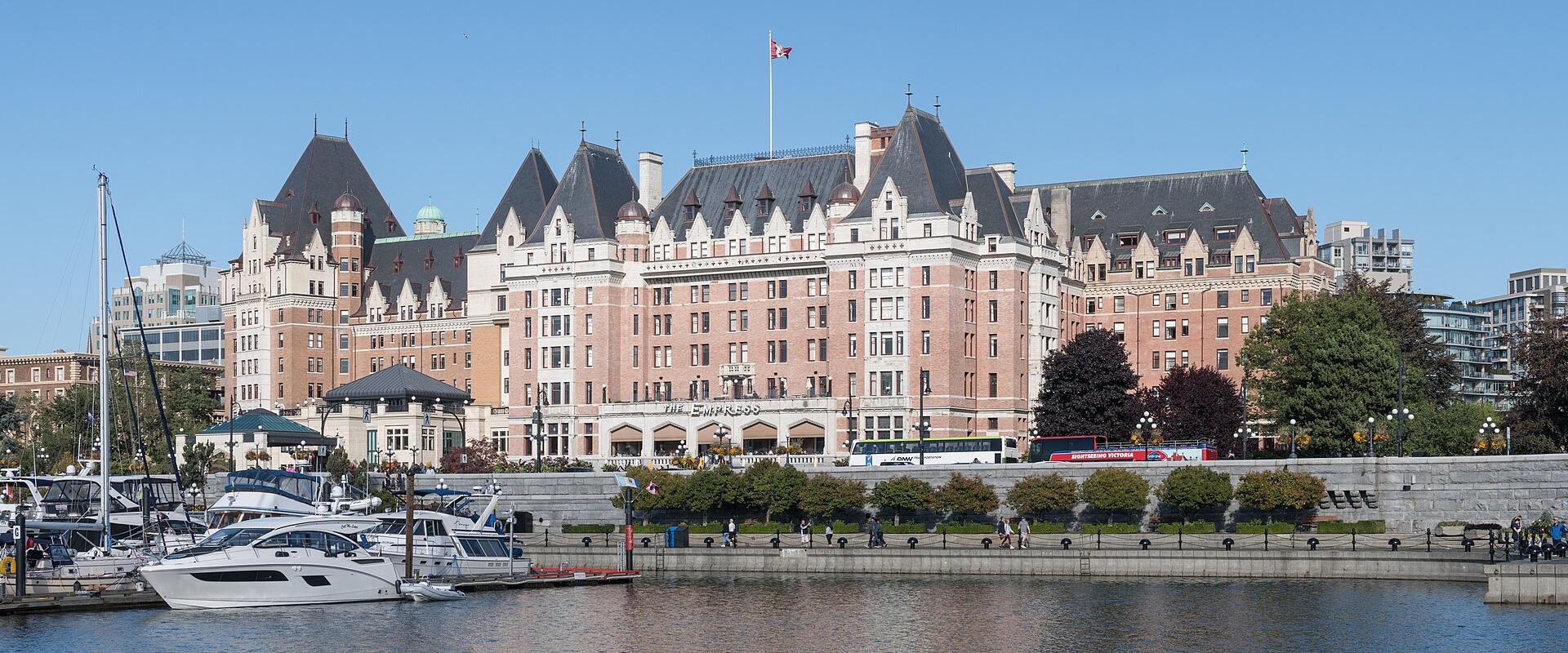 A large, historic pinkish-beige castle-style hotel with multiple turrets and a Canadian flag on the roof, situated on a waterfront promenade with docked boats and yachts in the foreground.