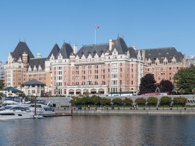 A large, historic pinkish-beige castle-style hotel with multiple turrets and a Canadian flag on the roof, situated on a waterfront promenade with docked boats and yachts in the foreground.