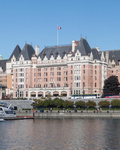 A large, historic pinkish-beige castle-style hotel with multiple turrets and a Canadian flag on the roof, situated on a waterfront promenade with docked boats and yachts in the foreground.